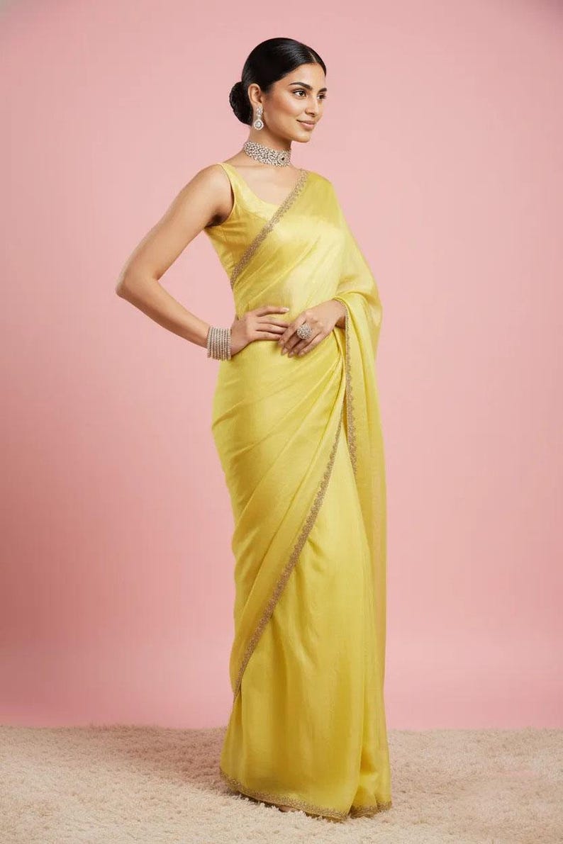 May include: A sleeveless, bright yellow sari featuring a subtle sheen and a delicate, thin embroidered border along the edges. The garment is paired with a matching sleeveless blouse, a sparkling silver-toned choker necklace, and matching bracelets.