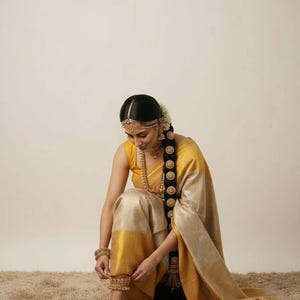 May include: A woman wearing a yellow and gold saree, adorned with gold jewelry, including a headpiece, earrings, necklace, bracelets, and anklets. Her long, dark hair is styled in a braid. The saree has a yellow border and a gold body.