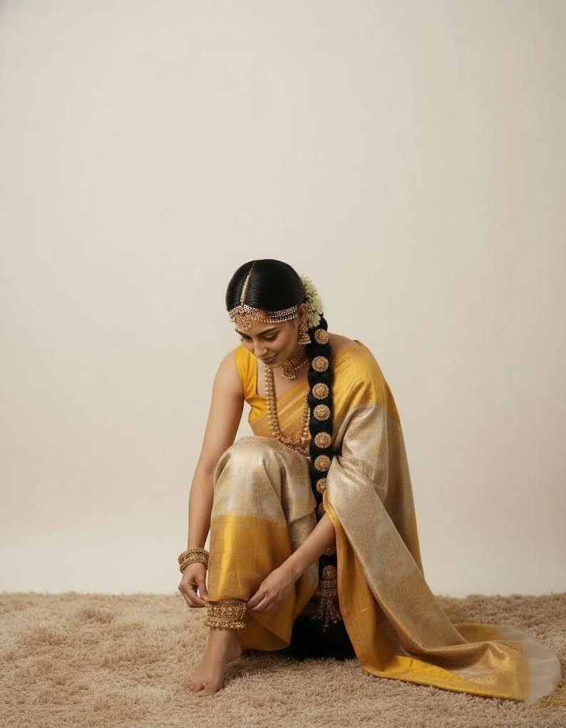 May include: A woman wearing a yellow and gold saree, adorned with gold jewelry, including a headpiece, earrings, necklace, bracelets, and anklets. Her long, dark hair is styled in a braid. The saree has a yellow border and a gold body.