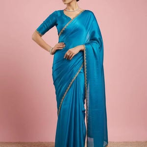 May include: A vibrant turquoise blue saree featuring a smooth, satin-like finish and a delicate gold embroidered border along the edges. The garment includes a matching short-sleeved blouse with a V-neckline and a long, flowing drape over the shoulder.