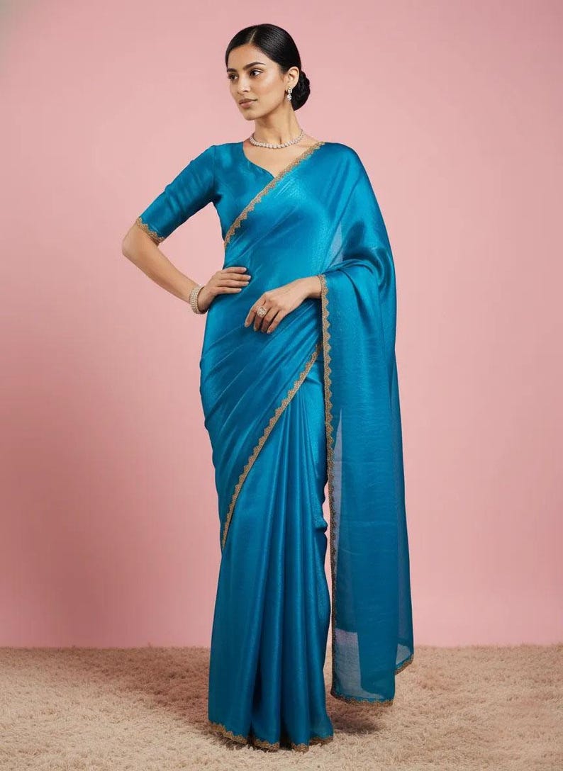 May include: A vibrant turquoise blue saree featuring a smooth, satin-like finish and a delicate gold embroidered border along the edges. The garment includes a matching short-sleeved blouse with a V-neckline and a long, flowing drape over the shoulder.