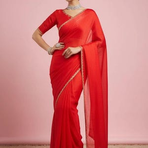 May include: A vibrant red saree featuring a V-neck blouse with short sleeves and a matching red skirt draped in a traditional style. The garment is accented with a delicate gold embroidered border along the edges of the fabric and the neckline.