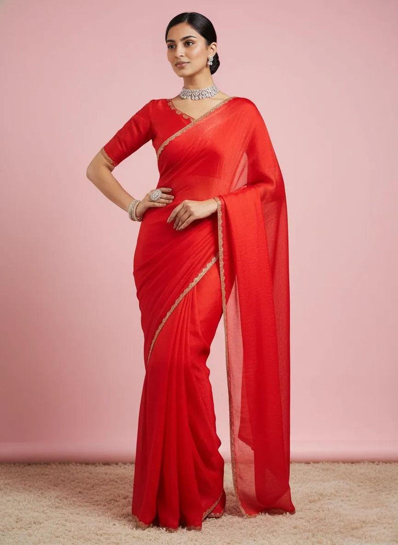 May include: A vibrant red saree featuring a V-neck blouse with short sleeves and a matching red skirt draped in a traditional style. The garment is accented with a delicate gold embroidered border along the edges of the fabric and the neckline.