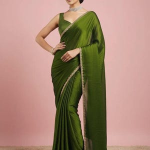 May include: An olive green saree featuring a sleeveless blouse with a square neckline and a matching skirt with a subtle sheen. The garment is adorned with a delicate, thin gold embroidered border along the edges, paired with a silver choker necklace.