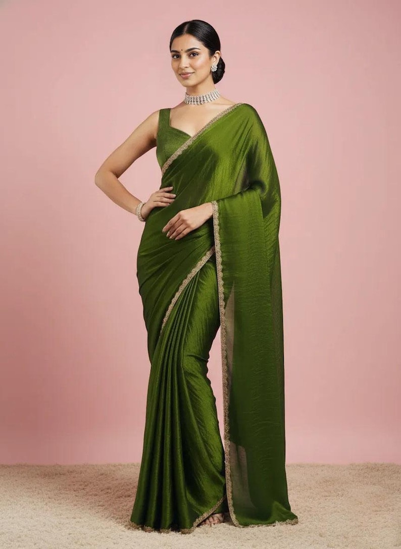 May include: An olive green saree featuring a sleeveless blouse with a square neckline and a matching skirt with a subtle sheen. The garment is adorned with a delicate, thin gold embroidered border along the edges, paired with a silver choker necklace.