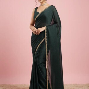 May include: A dark forest green saree featuring a sleeveless blouse and a matching drape with a subtle sheen. The edges of the fabric are finished with a delicate, scalloped gold metallic border that runs along the length of the garment.