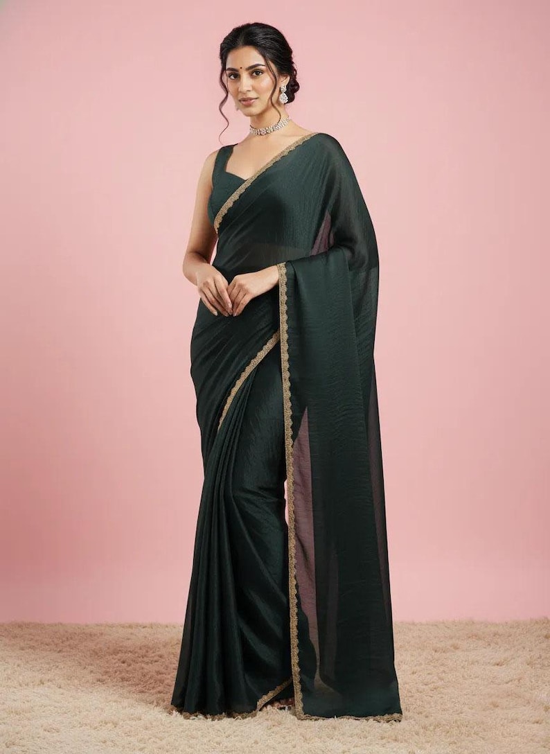 May include: A dark forest green saree featuring a sleeveless blouse and a matching drape with a subtle sheen. The edges of the fabric are finished with a delicate, scalloped gold metallic border that runs along the length of the garment.