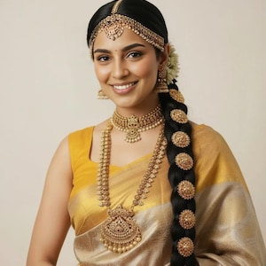 May include: A woman wearing traditional Indian gold jewelry, including a necklace, earrings, and headpiece. Her black hair is styled in a braid adorned with gold ornaments. She is wearing a yellow and gold sari.