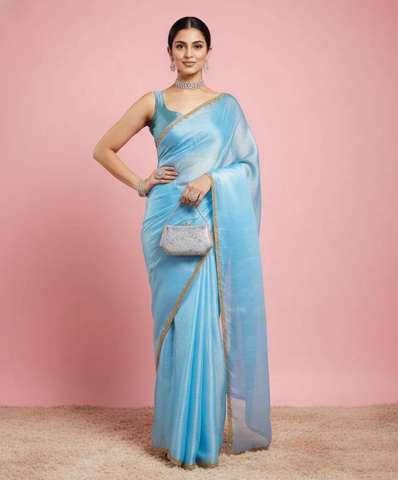 May include: A light blue saree with a subtle shimmer and a thin gold border, paired with a matching sleeveless blouse. The ensemble is accessorised with a silver-toned, rectangular, embellished clutch bag, a choker necklace, and bracelets.