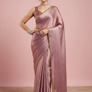 May include: A mauve-coloured saree with a subtle metallic sheen and a sleeveless blouse. The garment features a delicate gold-toned border along the edges. The ensemble is accessorised with a silver-toned choker necklace, matching earrings, and a bracelet.
