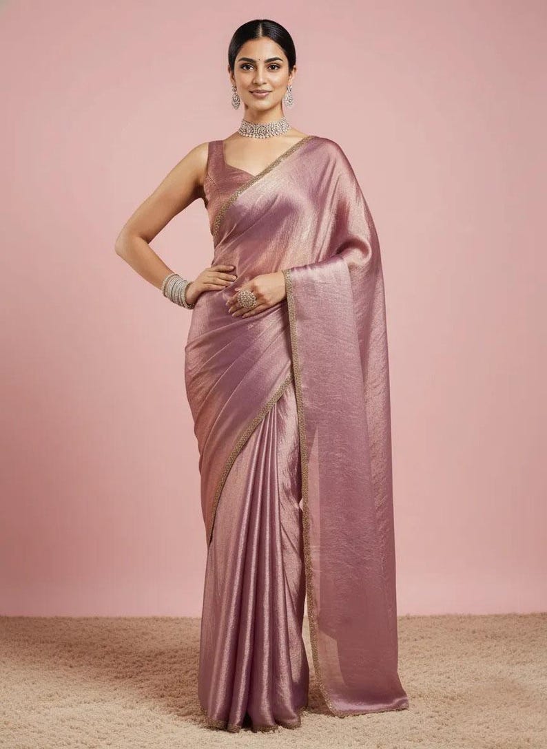 May include: A mauve-coloured saree with a subtle metallic sheen and a sleeveless blouse. The garment features a delicate gold-toned border along the edges. The ensemble is accessorised with a silver-toned choker necklace, matching earrings, and a bracelet.