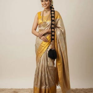 May include: A woman wearing a gold and beige sari with gold trim and a matching yellow blouse. She is adorned with gold jewelry, including a necklace, earrings, and headpiece. Her long, dark hair is styled in a braid with gold accents.
