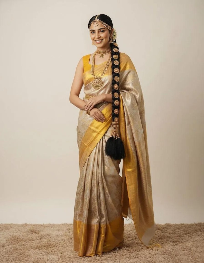 May include: A woman wearing a gold and beige sari with gold trim and a matching yellow blouse. She is adorned with gold jewelry, including a necklace, earrings, and headpiece. Her long, dark hair is styled in a braid with gold accents.