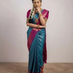 May include: A woman wearing a teal and magenta sari with gold trim. The sari is draped elegantly, with intricate patterns visible. She is adorned with gold jewelry, including a necklace, earrings, and bangles. Her hair is styled with a floral braid and a gold headpiece.