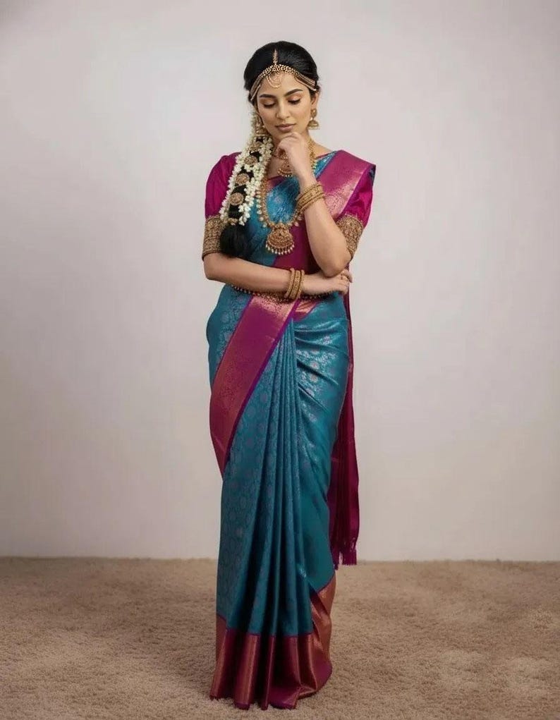 May include: A woman wearing a teal and magenta sari with gold trim. The sari is draped elegantly, with intricate patterns visible. She is adorned with gold jewelry, including a necklace, earrings, and bangles. Her hair is styled with a floral braid and a gold headpiece.