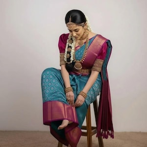 May include: A woman wearing a traditional Indian sari. The sari is teal with a patterned design and a magenta border. The blouse is magenta with gold detailing. She is adorned with gold jewelry, including a headpiece, earrings, and a long necklace.