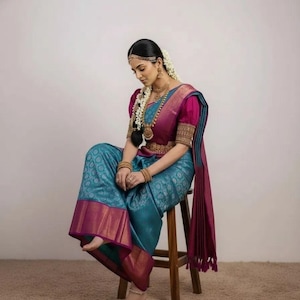 May include: A traditional teal and magenta sari with gold accents. The sari features a detailed pattern and is paired with a magenta blouse with puffed sleeves. The ensemble is accessorized with gold jewelry and a floral hairpiece. The person is seated on a wooden stool.