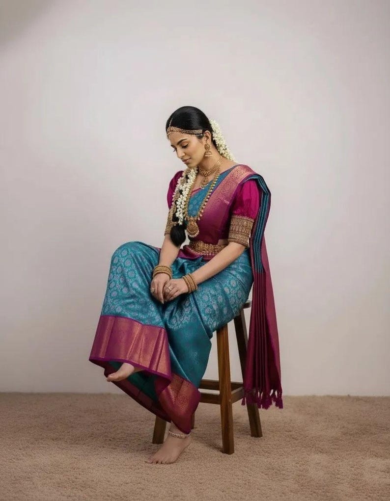 May include: A traditional teal and magenta sari with gold accents. The sari features a detailed pattern and is paired with a magenta blouse with puffed sleeves. The ensemble is accessorized with gold jewelry and a floral hairpiece. The person is seated on a wooden stool.