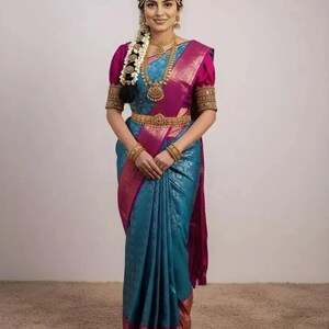 May include: A woman wearing a traditional Indian sari. The sari is teal with a magenta border and accents. The blouse is magenta with gold embroidered sleeves. Gold jewelry includes a necklace, earrings, and a belt. A floral hairpiece and headpiece complete the look.