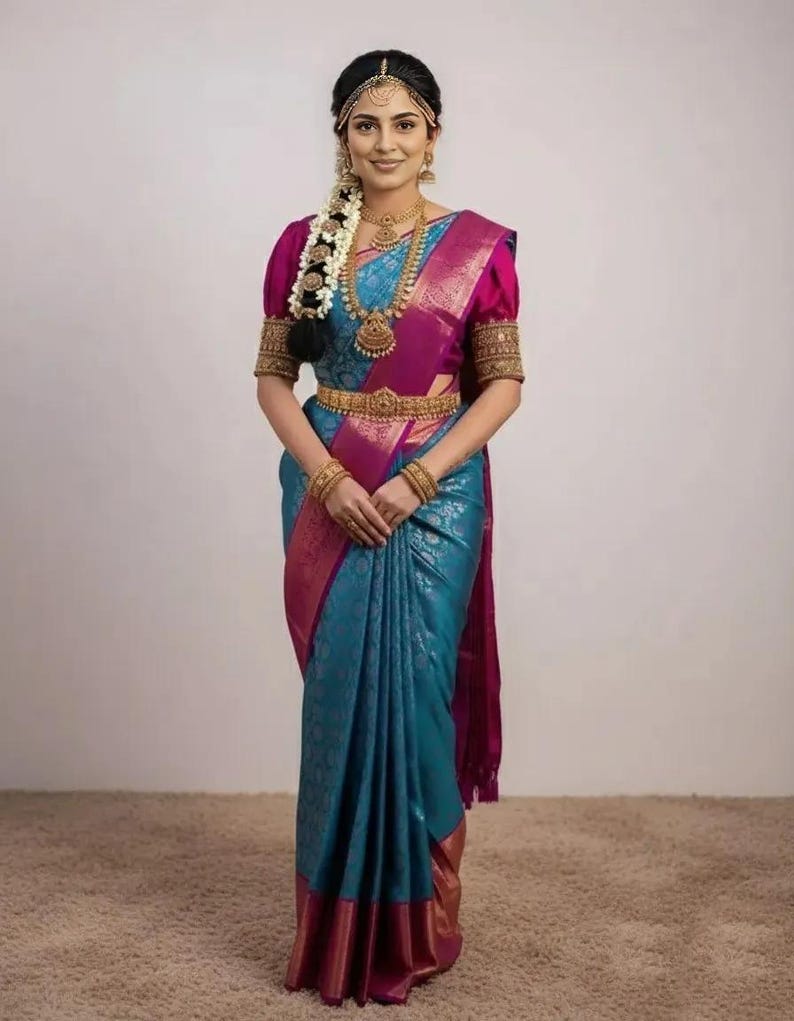 May include: A woman wearing a traditional Indian sari. The sari is teal with a magenta border and accents. The blouse is magenta with gold embroidered sleeves. Gold jewelry includes a necklace, earrings, and a belt. A floral hairpiece and headpiece complete the look.