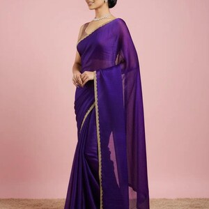 May include: A deep purple sari featuring a sheer, lightweight fabric with a subtle sheen. The garment is draped elegantly and finished with a delicate, scalloped gold-toned border along the edges, paired with a sleeveless matching blouse.