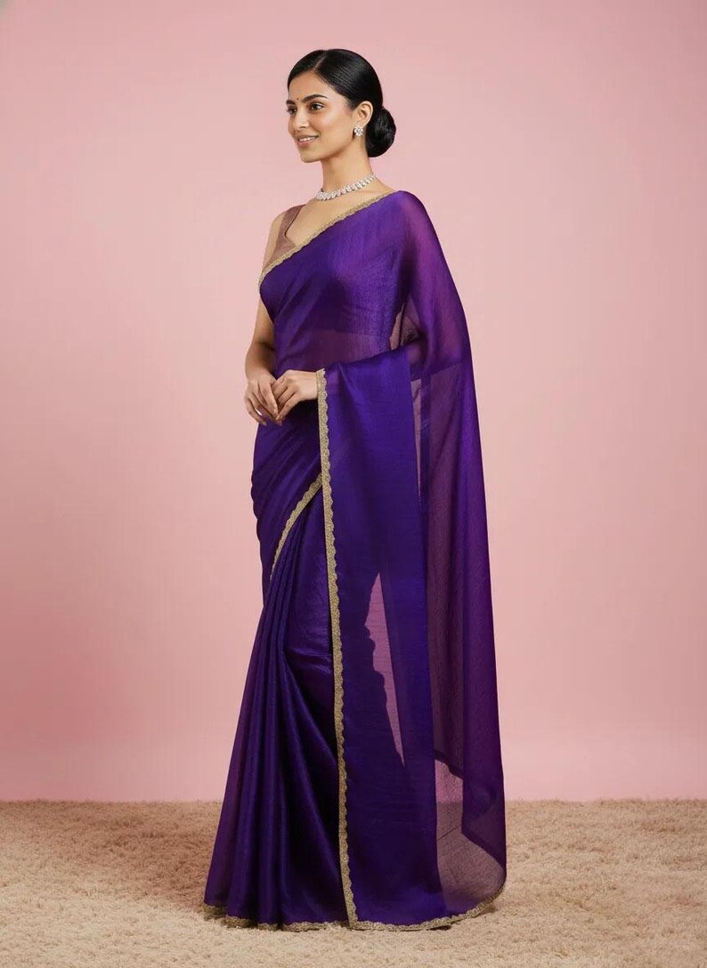 May include: A deep purple sari featuring a sheer, lightweight fabric with a subtle sheen. The garment is draped elegantly and finished with a delicate, scalloped gold-toned border along the edges, paired with a sleeveless matching blouse.