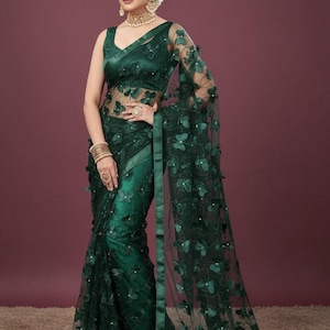 May include: A dark green saree with butterfly appliqués. The saree features a sheer overlay with embroidered butterflies and a matching sleeveless blouse. The model is wearing gold jewelry and a headpiece. The background is a solid maroon color.
