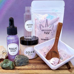 May include: A collection of bath products, including a bottle of "Magic Drops", a jar of "Color Fizz", and a bag of "Unicorn Fizz". The items are arranged on a wooden tray with decorative stones and a wooden spoon. A rainbow is in the background.