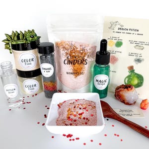 May include: A collection of fantasy-themed potion ingredients and accessories. Includes jars labeled "Color Fizz" and "Embers & Ash", a bottle of "Magic Drops", and a bag of "Glowing Cinders". A recipe card and a wooden spoon are also present.