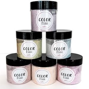 May include: A stack of six clear plastic jars with black lids, each filled with colorful bath fizz. Each jar has a white label with the words "COLOR Fizz" in a circular design. The colors of the fizz include pink, blue, green, and purple.