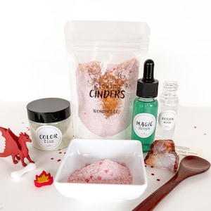 May include: A collection of fantasy-themed bath products, including a jar labeled "Color Fizz", a bag of "Glowing Cinders", and bottles of "Magic Drops" and "Potion Water". A red dragon figurine, a white bone, and a small flame are also included.