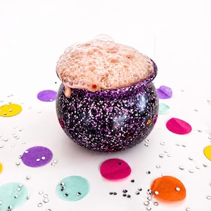 May include: A black and purple glitter-covered cauldron with a bubbling, brown foam overflowing from the top. Colorful confetti and silver glitter are scattered around the cauldron on a white background. A whimsical, festive image.