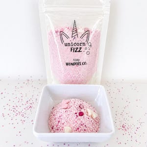 May include: A clear bag of pink Unicorn Fizz bath powder with a unicorn horn logo and the text "tiny Wonders Co." A white square bowl contains a mound of pink bath powder with decorative sprinkles.