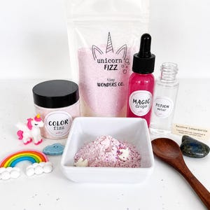 May include: A collection of colorful bath products and accessories. Includes a jar of "Color Fizz", a bag of "Unicorn Fizz", "Magic Drops", and "Potion Water". Also features a rainbow, a unicorn figurine, and a wooden spoon.