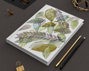 May include: A white journal with a watercolor illustration of green, yellow, and purple leaves. Gold and black pencils and gold binder clips are arranged around the journal on a dark gray surface.