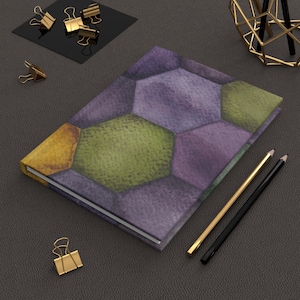 May include: A hardcover journal with a geometric pattern in shades of purple, green, and gold. The cover has a textured appearance, and the journal is accompanied by gold and black pencils and gold binder clips.