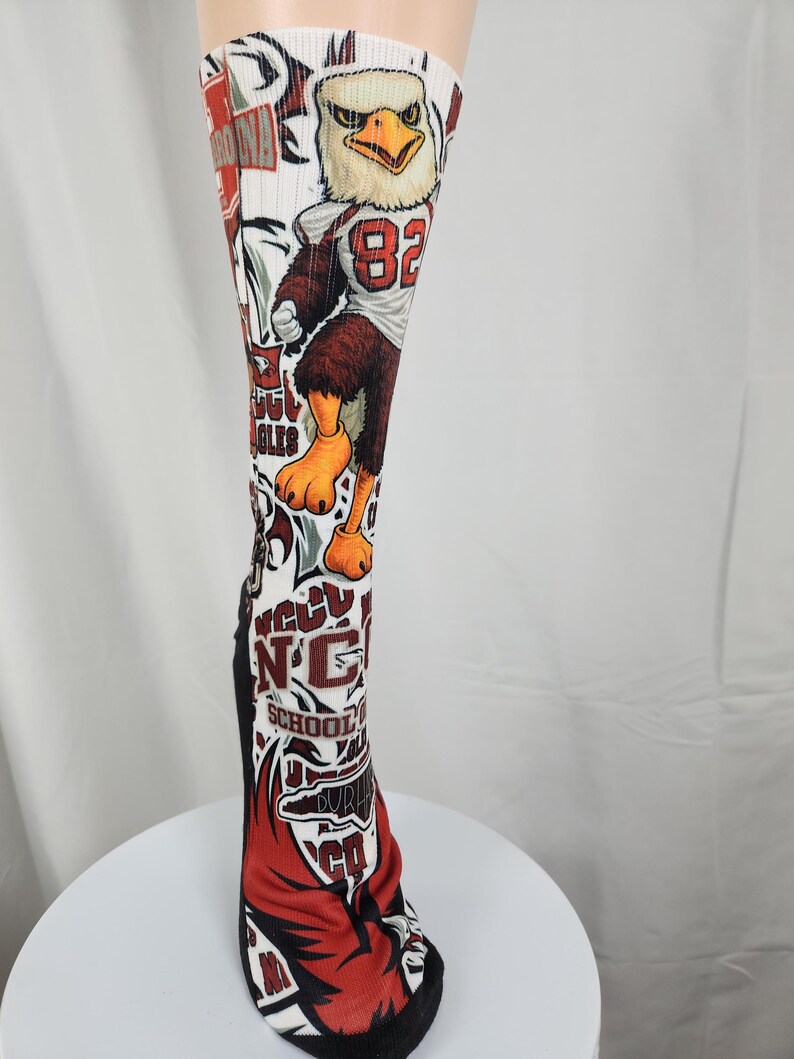 May include: A white knee-high athletic sock featuring a graphic print of an eagle mascot wearing a jersey with the number 82. The design includes red and maroon accents, text reading NCCU SCHOOL OF LAW, and additional sports-themed graphics.