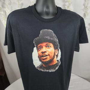 Pu&ograve; includere: T-shirt nero girocollo con un ritratto di un uomo con capelli scuri e barba. Il ritratto &egrave; in stile pittorico, con toni caldi e uno sfondo bianco. La t-shirt &egrave; esposta su un manichino nero.