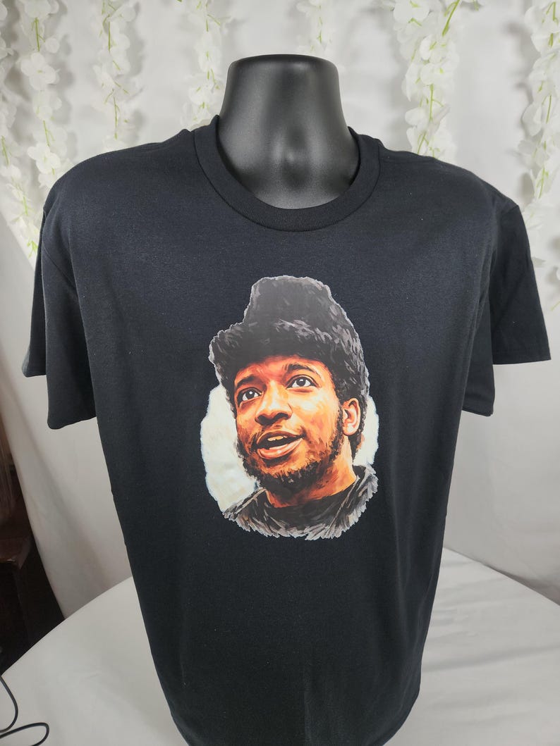 Pu&ograve; includere: T-shirt nero girocollo con un ritratto di un uomo con capelli scuri e barba. Il ritratto &egrave; in stile pittorico, con toni caldi e uno sfondo bianco. La t-shirt &egrave; esposta su un manichino nero.