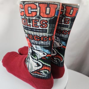 May include: A pair of red and multi-colored socks. The socks feature the text "CCU EAGLES" in orange and white, with a graphic of an eagle. The socks are displayed on a white surface.