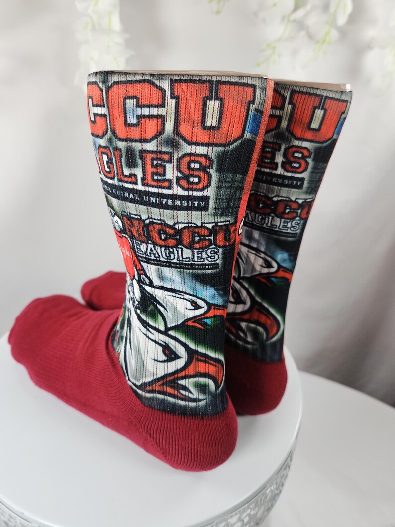 May include: A pair of red and multi-colored socks. The socks feature the text "CCU EAGLES" in orange and white, with a graphic of an eagle. The socks are displayed on a white surface.