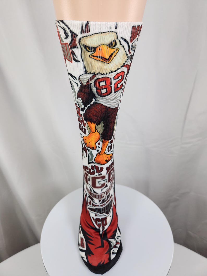 May include: A white knee-high athletic sock featuring a graphic of a cartoon eagle mascot wearing a jersey with the number 82. The sock includes red and black accents, with text elements reading NCCU, SCHOOL OF LAW, and DURHAM printed along the leg.