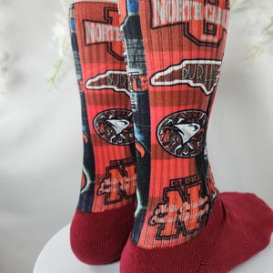 May include: Pair of red and black crew socks with the words "NORTH CAROLINA" and "DURHAM" printed on them. The socks also feature a logo with a bird and the text "EST 1910".