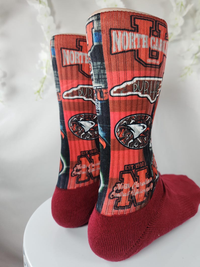 May include: Pair of red and black crew socks with the words "NORTH CAROLINA" and "DURHAM" printed on them. The socks also feature a logo with a bird and the text "EST 1910".