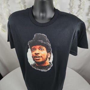 Pu&ograve; includere: T-shirt nero con un ritratto di un uomo con capelli scuri e barba. Il ritratto &egrave; in un cerchio bianco. La t-shirt &egrave; esposta su un manichino nero.