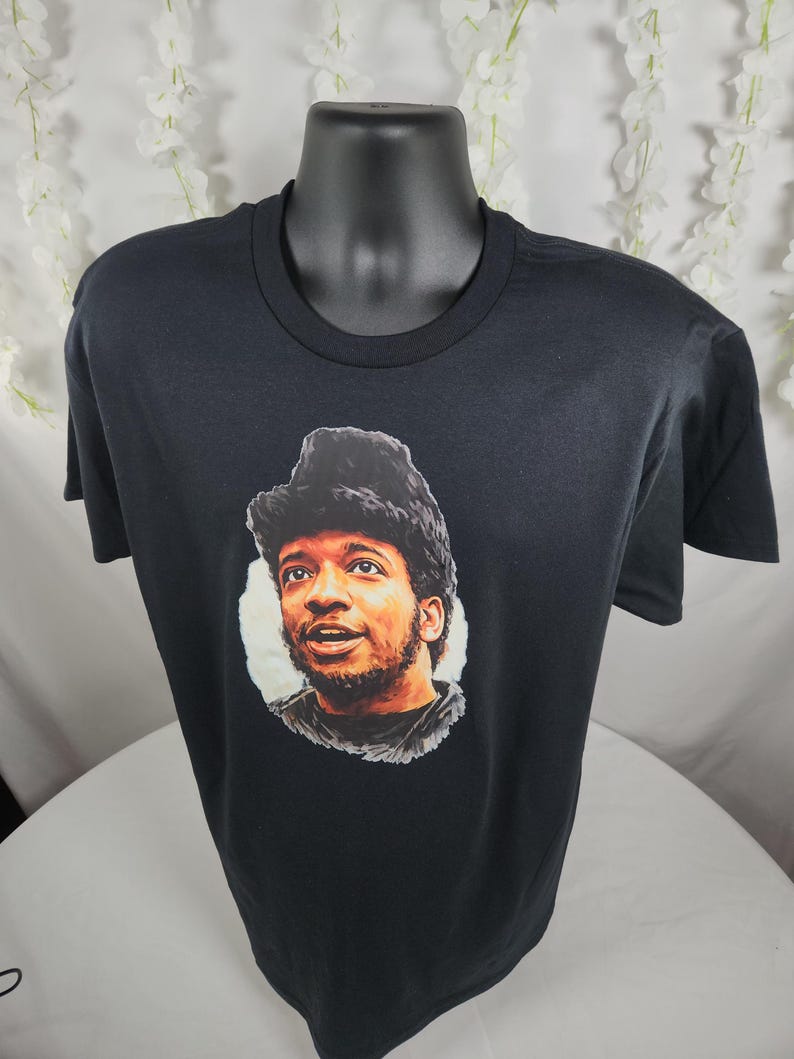 Pu&ograve; includere: T-shirt nero con un ritratto di un uomo con capelli scuri e barba. Il ritratto &egrave; in un cerchio bianco. La t-shirt &egrave; esposta su un manichino nero.