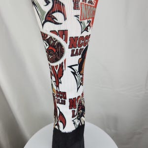 May include: A white crew sock featuring an all-over print of red and black eagle graphics and the text NCCU EAGLES. The sock has a solid black heel and toe section, with various repeating logos and team-related imagery displayed across the white fabric.