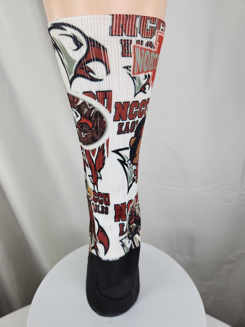 May include: A white crew sock featuring an all-over print of red and black eagle graphics and the text NCCU EAGLES. The sock has a solid black heel and toe section, with various repeating logos and team-related imagery displayed across the white fabric.