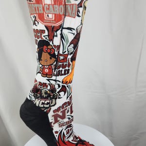 May include: A white crew sock featuring a repeating graphic print of North Carolina Central University logos, eagle mascots, and a cartoon character with a red bow. The sock has a solid black heel and toe section with ribbed fabric at the top cuff.