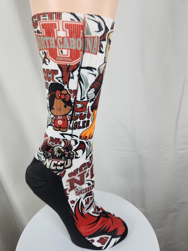 May include: A white crew sock featuring a repeating graphic print of North Carolina Central University logos, eagle mascots, and a cartoon character with a red bow. The sock has a solid black heel and toe section with ribbed fabric at the top cuff.